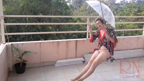 A sexy babe swings on a hanging swing, staying dry under an umbrella from the rain.