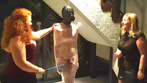 Two gorgeous femdoms dominate a plump male slave wrapped in cling film, giving him CBT with a riding crop!